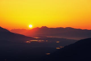 The sun is about to set behind a mountain chain, colouring the sky golden and the mountain ridges red. There is flat land with several lakes coloured gold and red in the mid-section of the photo. Fog coloured slightly blue whiffs over the earth.