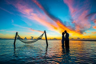 A couple in silhouette kissing while standing in thigh-deep water with a spectacular sunset in the background.