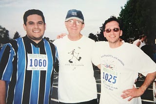 Three men standing side by side at an outdoor event, smiling at the camera. The man on the left wears a blue and black striped sports jersey with a race number ‘1060,’ and the man on the right wears sunglasses and a white shirt with a race number ‘1025.’ The man in the center wears a white T-shirt with a hiking graphic and a ‘Grand Canyon’ hat.