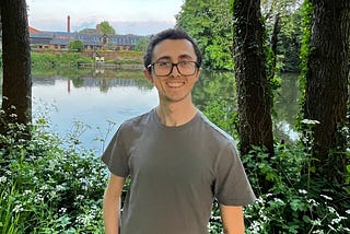 A man smiling in front of a body of water with trees lining the riverbank.