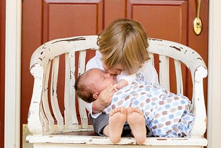 A boy sitting on a chair holding and kissing a baby.