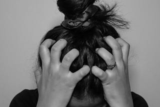 A black-and-white portrait of a person with their head down and their hands clawing at their hair, looking extremely tense and frustrated.