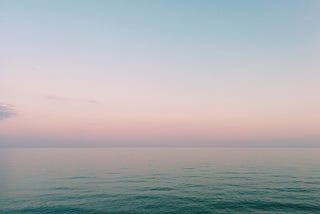 A beach at sunrise, with pastel hues in the sky and on the water blurring the transition between them.