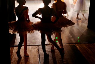 Young girls in tutus backstage at a rehearsal, watching ballerinas practicing on stage.