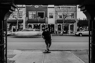 A black and white photograph of a man standing on the sidewalk, waiting to cross a street. The background features a row of small shops and buildings. A car is seen driving by in a blur, capturing motion, while the man remains still. The scene appears to be from a small town or suburban area.