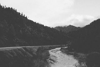 Black and white photo of a cloudy landscape. It follows a road, with trees on one side and rocks and a river on the other. There might be a mountain in the distance under the clouds.