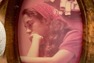 Framed faded photo of a young woman with long dark hair in a red kerchief with her chin in her hand.