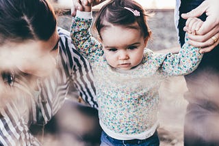 An image of a little girl around one or two years old being supported to stand up by her parents.