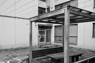 An ugly pavilion in a backyard made from broken concrete plates. In the background is a broken glass door secured with tape.