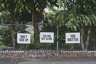 A chain-link fence with three signs that read ‘DON’T GIVE UP,’ ‘YOU ARE NOT ALONE,’ and ‘YOU MATTER,’ surrounded by greenery and trees in a peaceful outdoor setting.