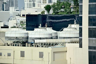 View of rooftop cooling towers and HVAC systems on top of a building in a cityscape, with high-rise buildings and trees visible in the background.