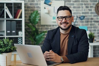 Male manager sitting at a desk in a modern office looking confident and smiling. | Photo by Vitaly Gariev on Unsplash