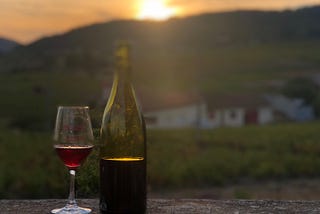 A bottle and glass of red wine with vineyards and sunset in the background