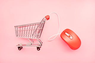 A small shopping cart and red computer mouse on pink background, symbolizing online shopping.