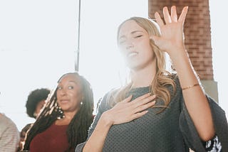 Blond lady in church with eyes closed and hand in air, in a religious trance, bu the lady behind her is looking at her as if she is crazy.