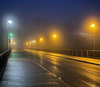 A deserted, foggy, wet road looks ominous at night with the only sign of life being the amber and white street lights illuminating the road.