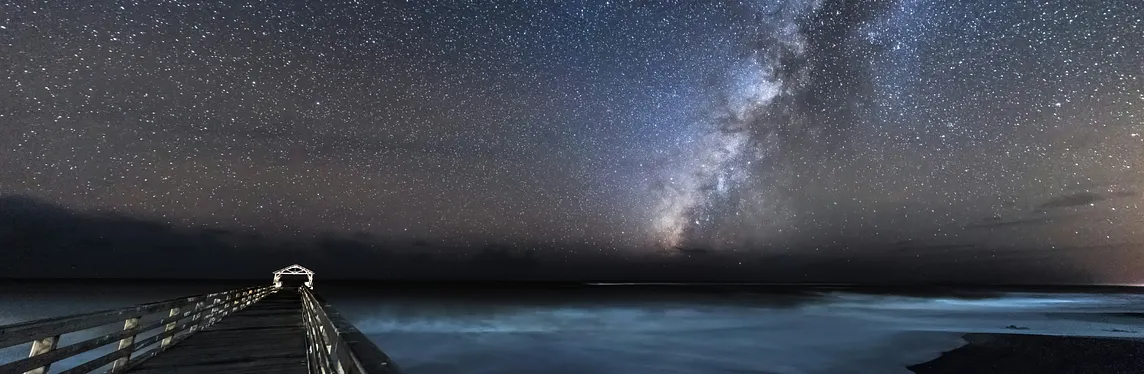 The Milky Way and stars dominate the night sky above a lake, with a pier in the foreground.