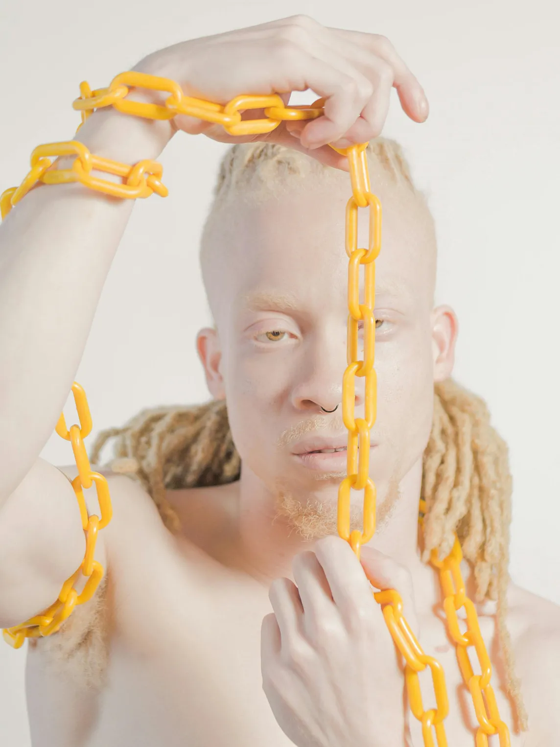 A close-up image of a man with white eyebrows and golden dreadlocks posing with a yellow chain wrapped around his arms and body. The backdrop is white.