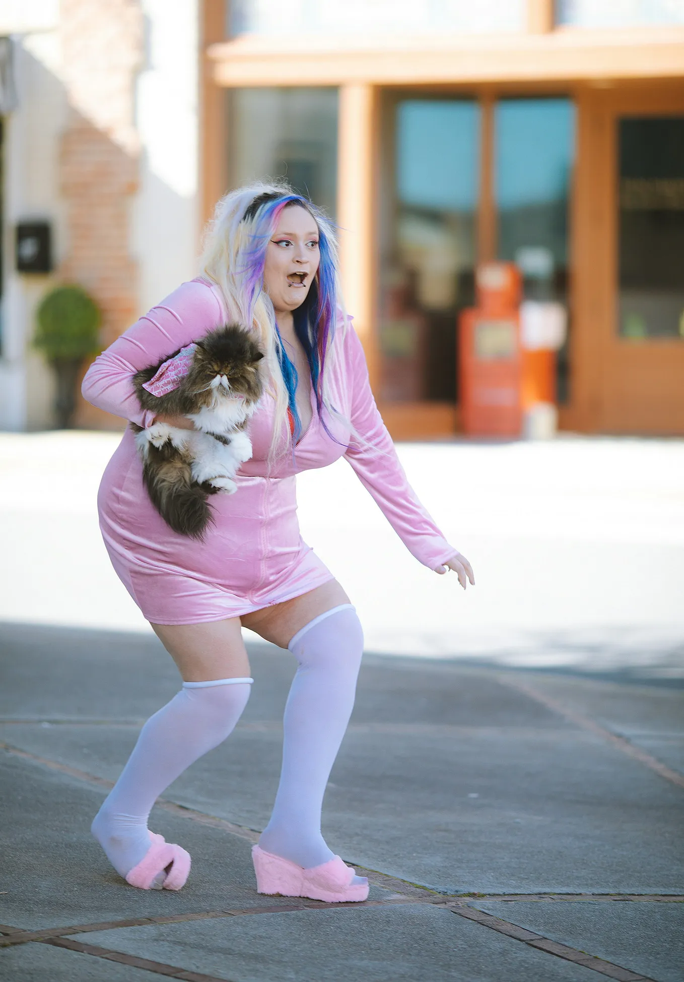 a woman with multicolored hair and a pink outfit is holding a fluffy cat and walking on the street