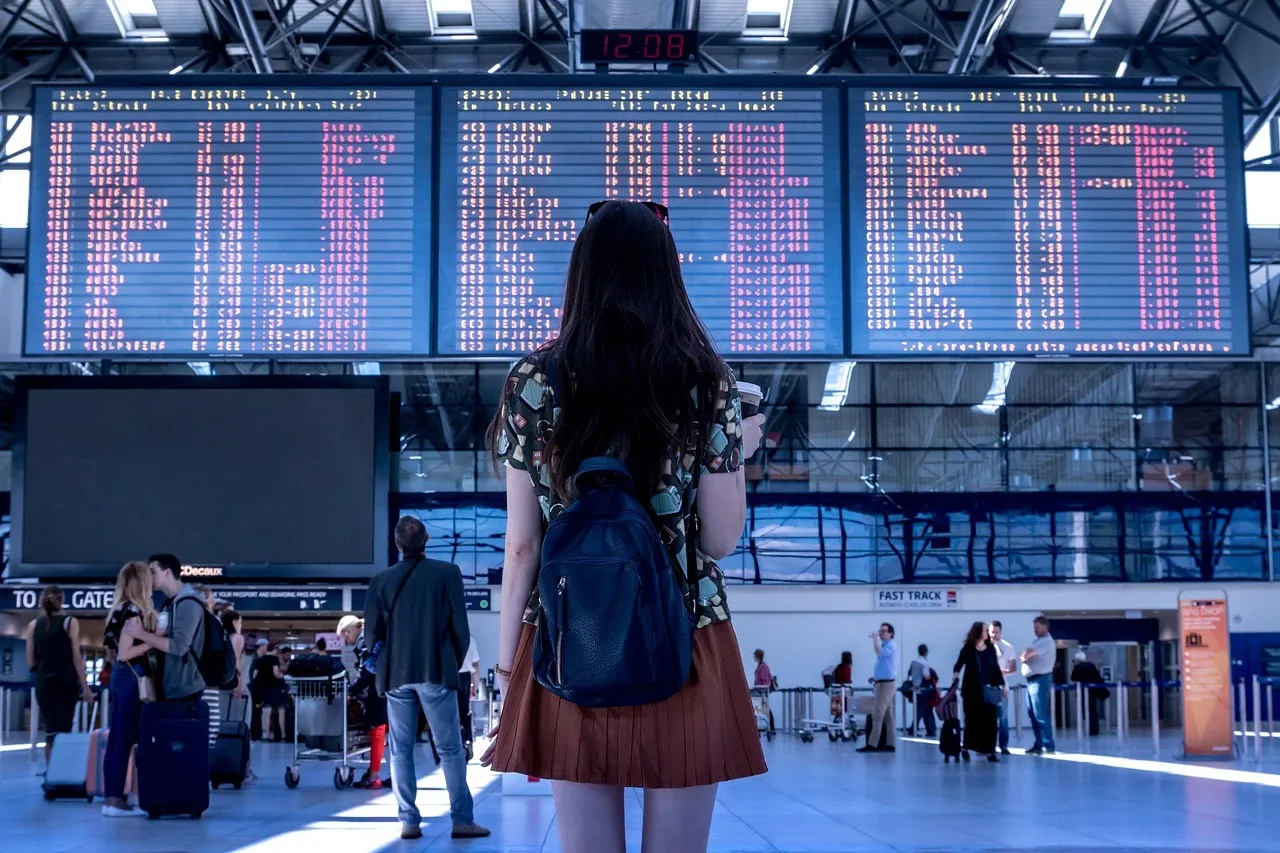 A woman with long black hair wearing a brown pleated skirt and gazing at the flight board at an airport.