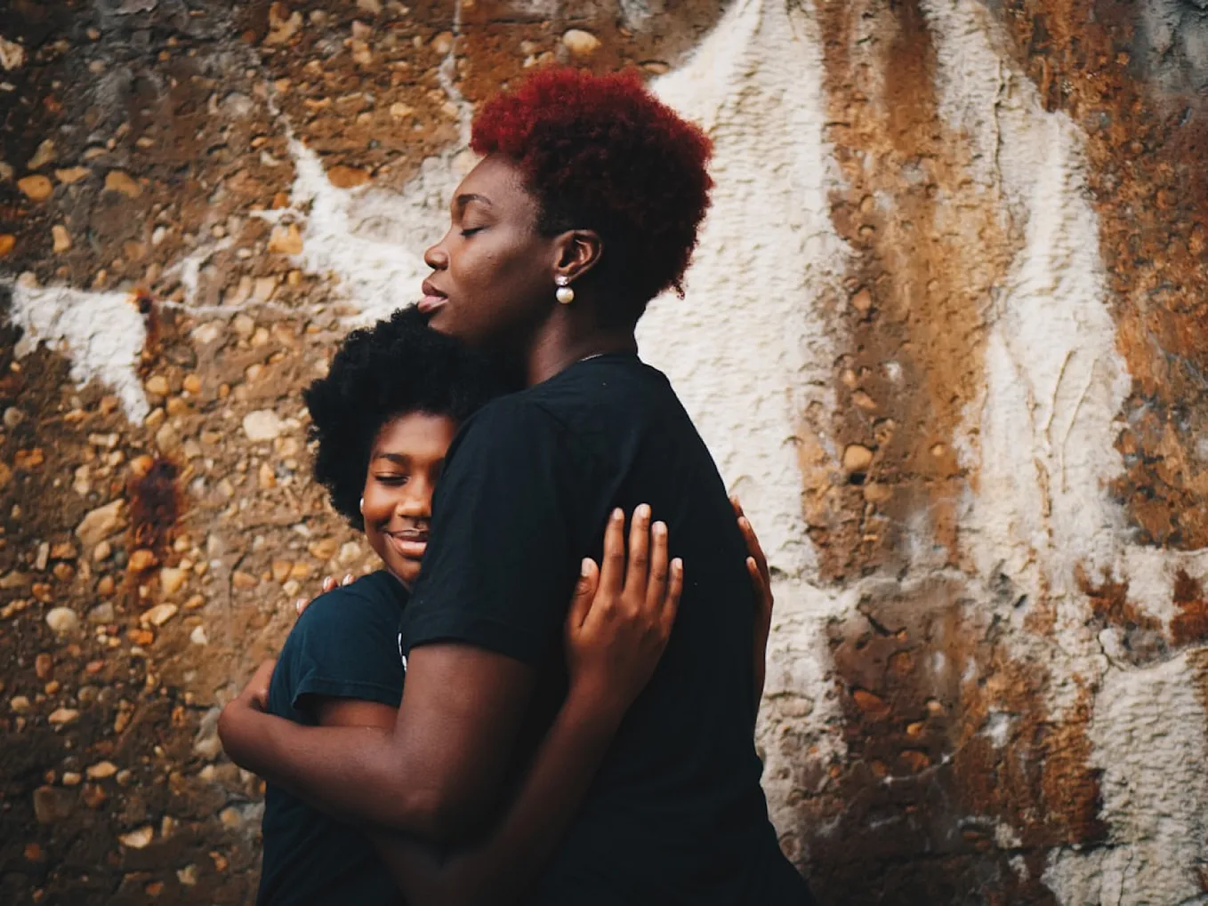 A black woman hugs a black girl. Both have their eyes closed as they embrace while standing in front of a wall.