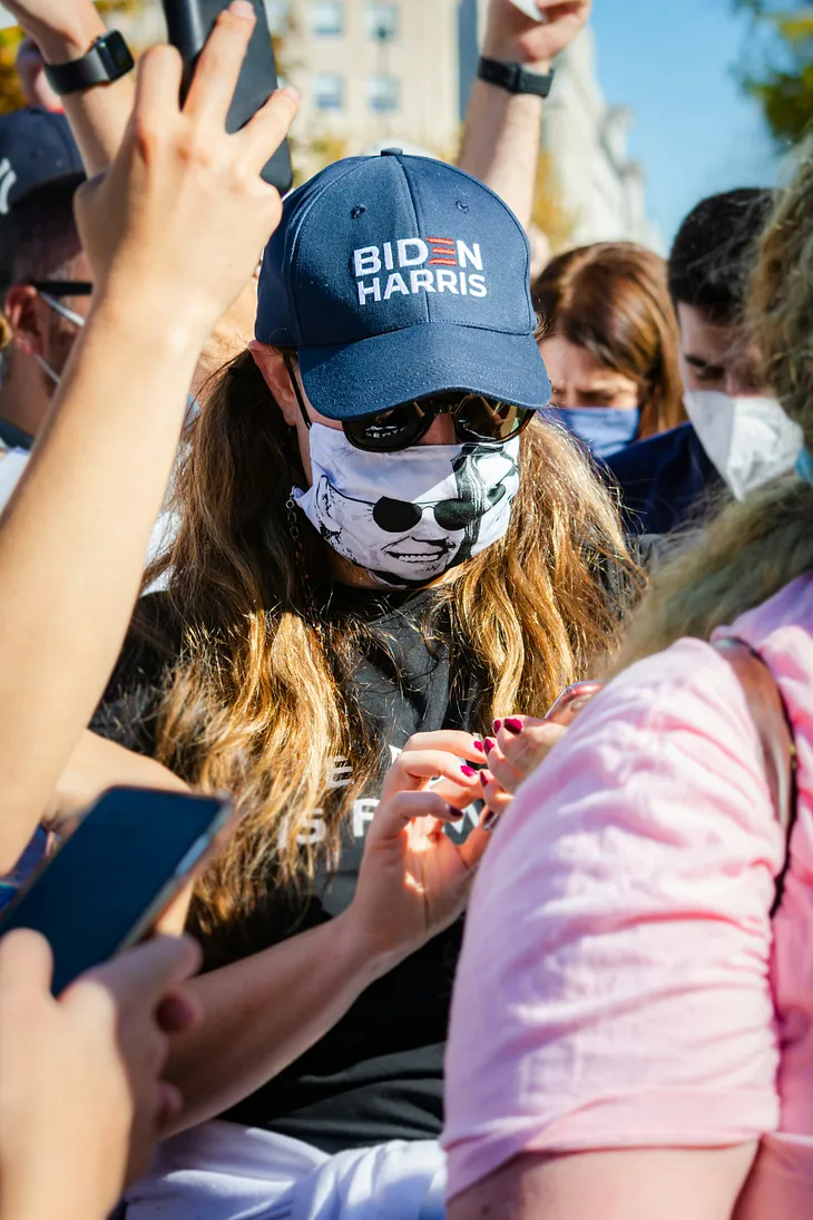 A person wearing a “BIDEN HARRIS” cap and a face mask with a graphic of a person wearing sunglasses is surrounded by other people. The person is holding a smartphone and appears to be looking at the screen. The crowd around them is also engaged with their phones. The person has long hair and is wearing sunglasses. The background is slightly blurred, emphasizing the central figure.