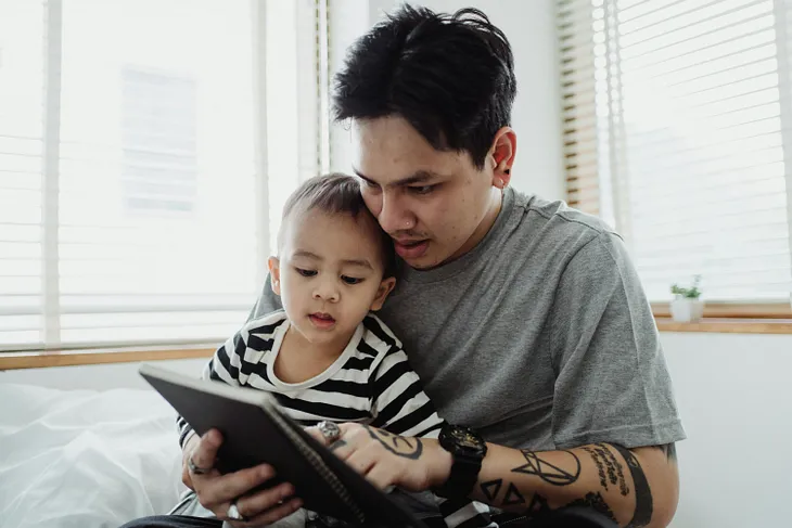 Very young son is sitting on his father’s lap while his father shows him a notebook.