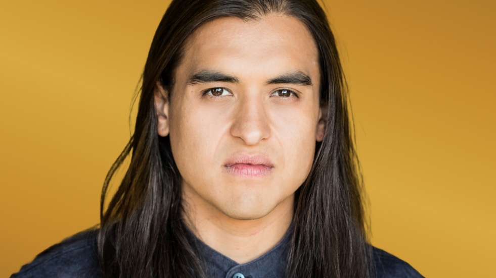 A young Cree man with dark eyes and long dark hair cascading around his shoulders.