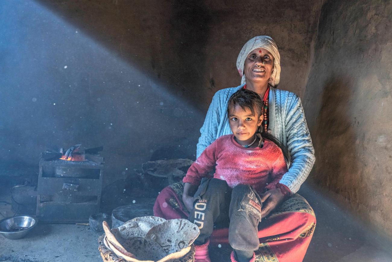 This is a photo clicked on a rural area kitchen where a girl is sitting on her mother's lap with some chapatis in front of her. 