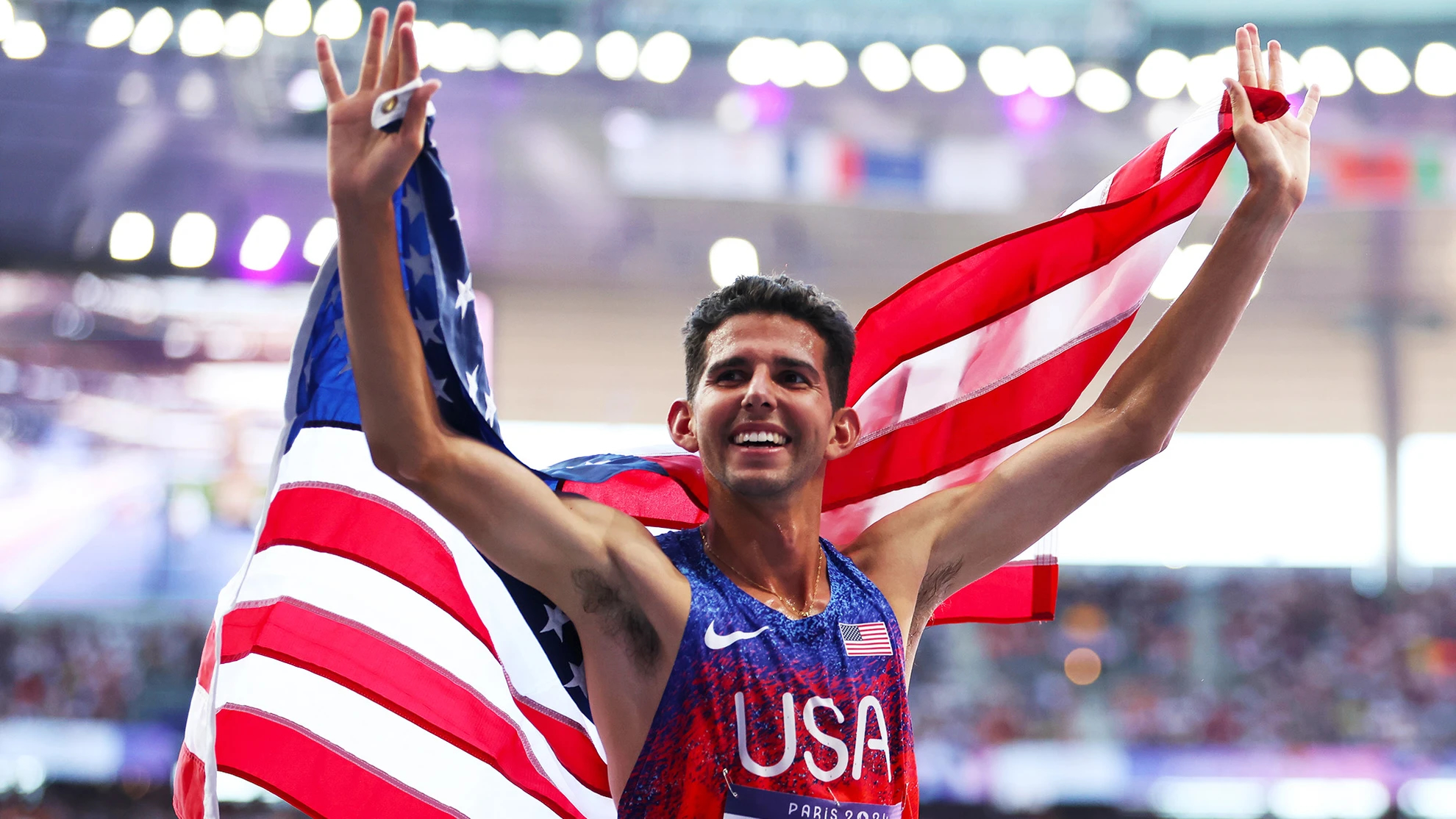 Track & field medalist Grant Fisher stands triumphantly, partially wrapped in an American flag.