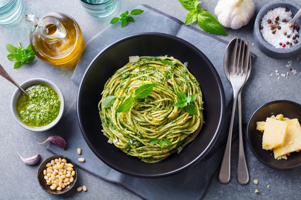 Top view of spaghetti pasta with pesto sauce and fresh basil leaves in a black bowl on a grey background