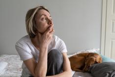 Middle-aged blonde woman in nightwear, sitting on bed with hand on chin, looking in distance with a depressed expression