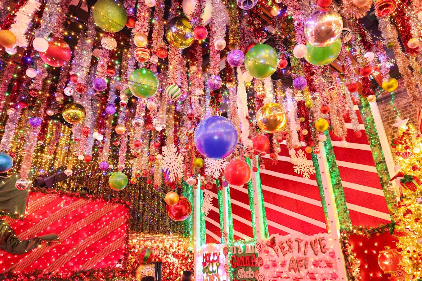 An extravagantly decorated room with a ceiling covered in colorful ornaments, tinsel, and holiday lights in various sizes and shades. The festive atmosphere is amplified by bright candy-cane-striped walls, oversized snowflakes, and signs reading "Festive AF!"