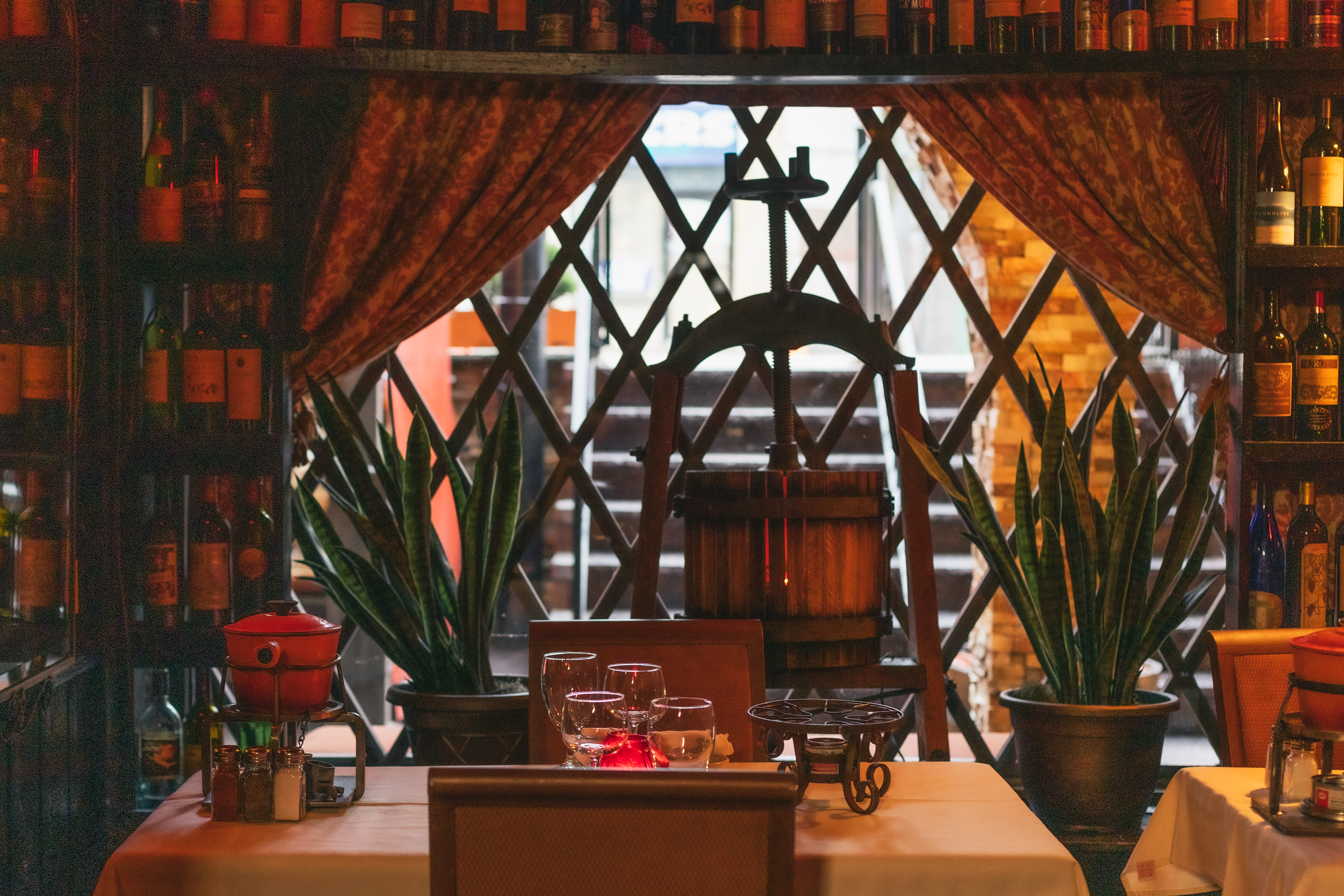 a cozy dining area with a rustic and intimate ambiance. The centerpiece is a wine press framed by lattice-style windows and rich drapery. Shelves filled with wine bottles and potted plants enhance the atmosphere, while a table is set with glassware and a red fondue pot, evoking a warm and inviting scene.