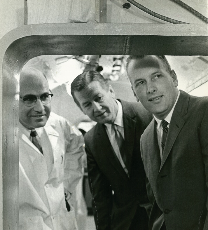 Three people in 1960s American dress look inside a large metal box