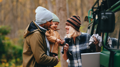 Smiling father, mother, and toddler outside of their van, bare autumn leaves in the background