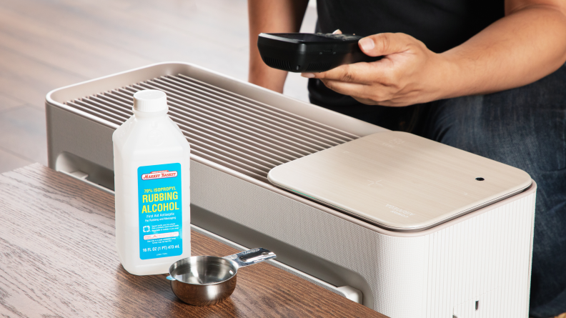 A small cup of rubbing alcohol sits next to a bottle of rubbing alcohol on a desk; next to the desk a hand holds an air quality monitor over an air purifier.