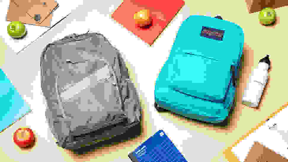 An L.L. Bean backpack and JanSport backpack laying on a table surrounded by school supplies and apples.