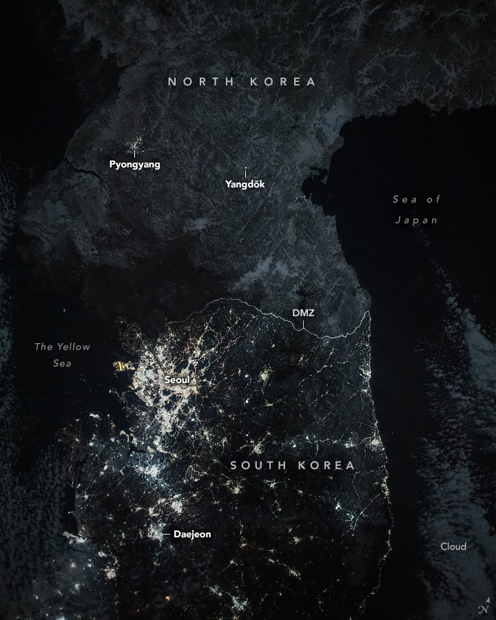 A night image of the Korean Peninsula shows the Sea of Japan, with cloud coverage in the lower right, and the Yellow Sea on the left of the peninsula. The land highlights capital cities of Pyongyang in the North, and Seoul in the South. There is some illumination visible from Pyongyang, while Seoul is brightly illuminated. The city of Daejeon is also labelled and well illuminated. In the North, Yangdŏk is labelled and appears to have some night illumination as well. The DMZ border between the two countries is highlighted.