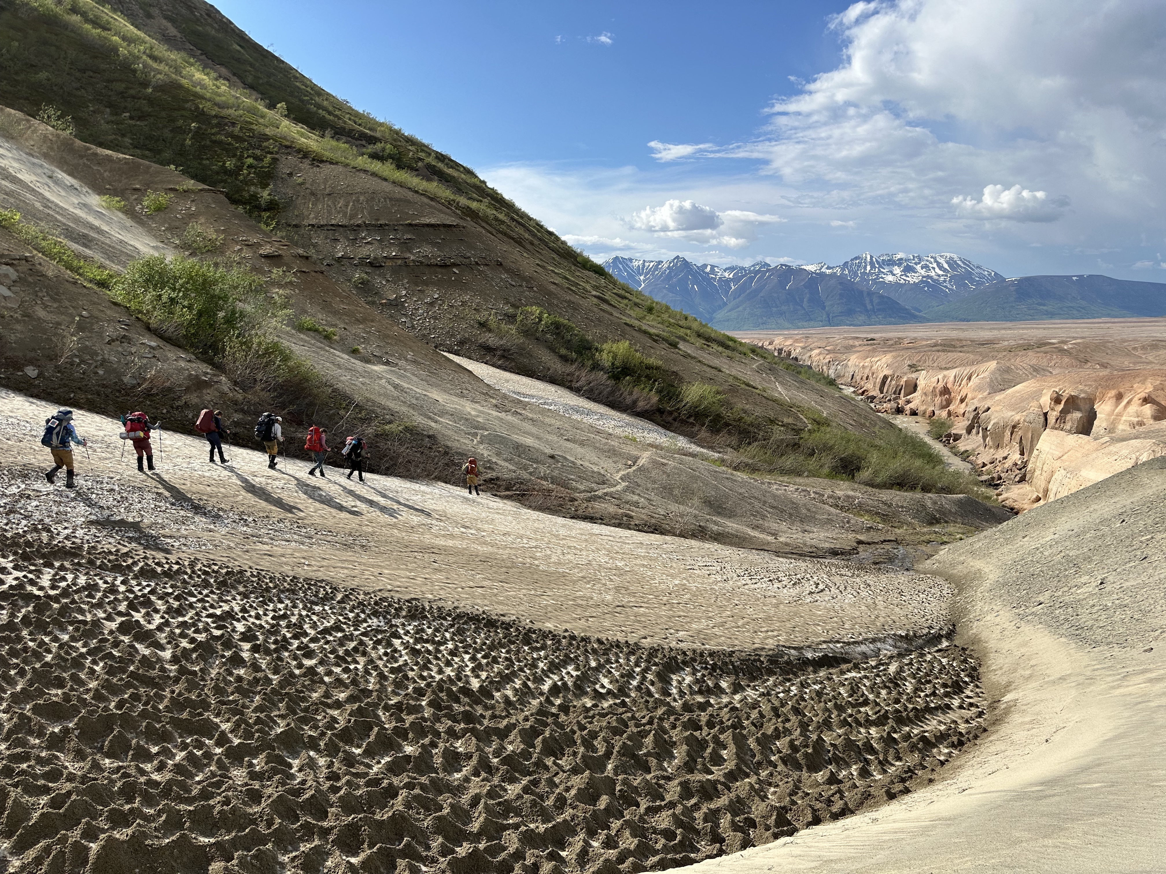 Seven people, with large backpacks, hiking down a hill of lumpy snow dusted with beige volcanic ash. Behind them is a steep wall of dirt with streaks of fresh green shrubbery. The people appear tiny against the landscape and are all in the left half of the image. On the right are overlapping views of three distinct geological formations: a light-colored slope in the foreground, a tan and orange river gorge in the middle ground, and snow-capped mountains in the background, under a partly cloudy sky.