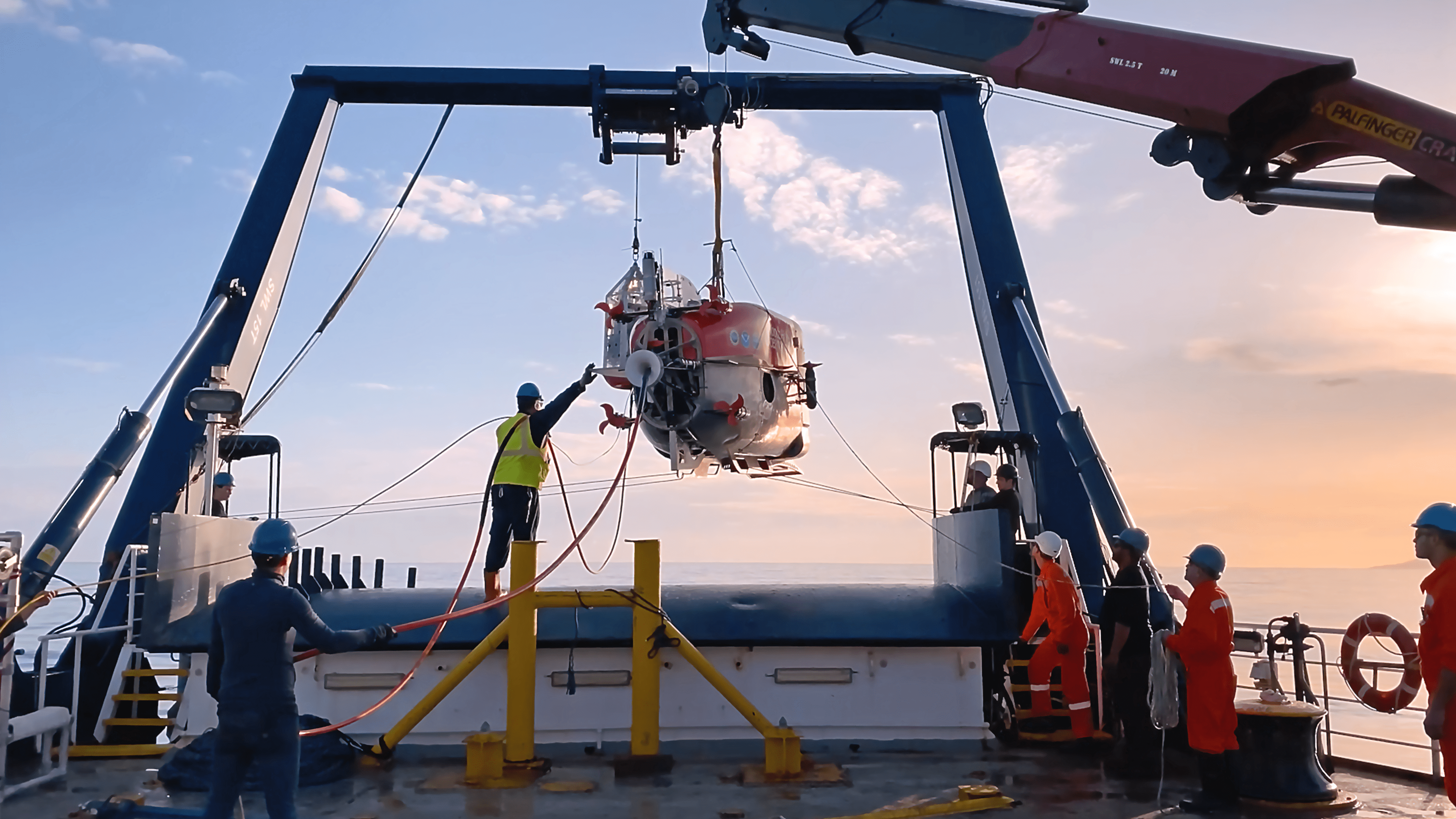 On the deck of a ship, a large A-frame style crane is lifting a submersible research vehicle into the air, as crew members hold taglines connecting to the vehicle, preparing to deploy it into the ocean.