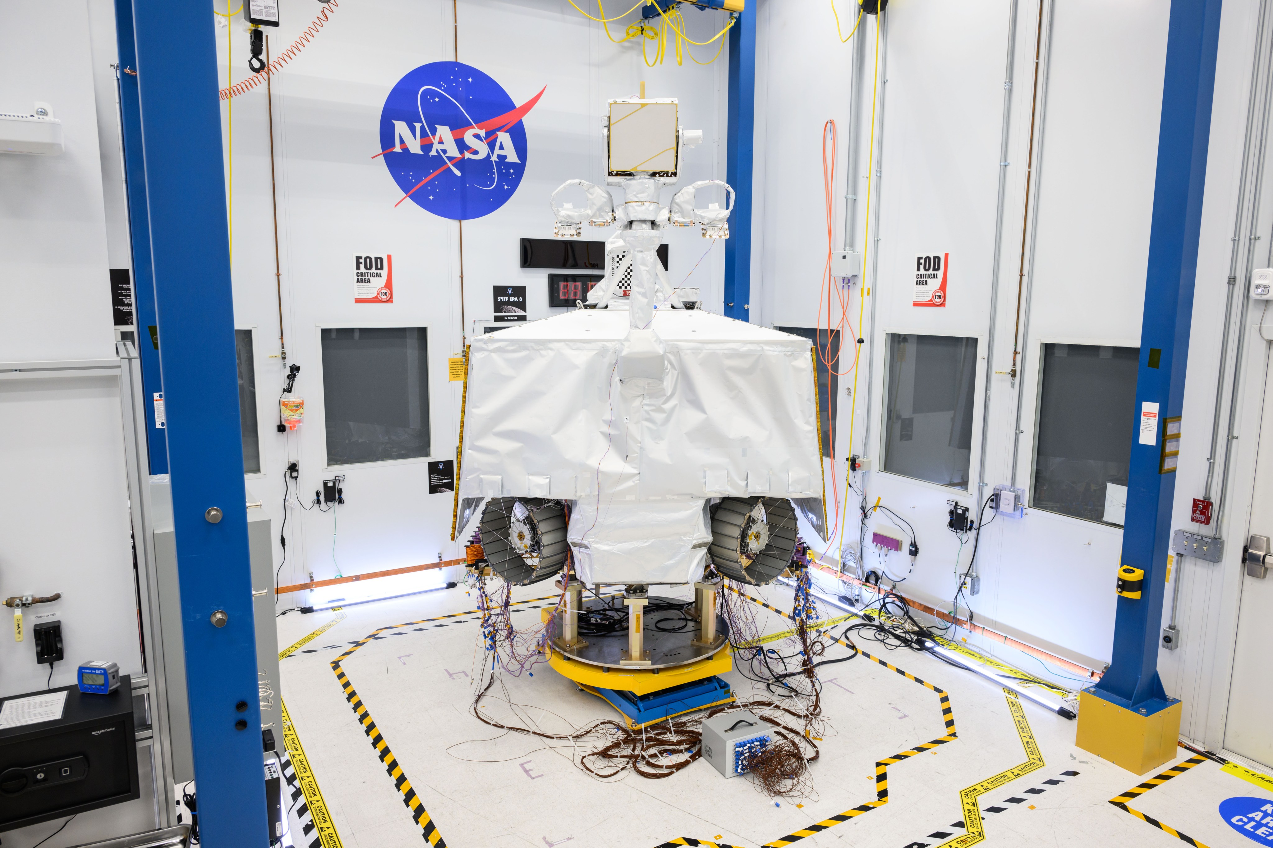 NASA’s VIPER robotic Moon rover is shown in a clean room at NASA’s Johnson Space Center in Houston. Two of its wheels are visible in this image. It is shown on a white floor in front of a white wall next to the NASA "meatball" logo. On the the floor there is yellow-and-black tape surrounding the rover.