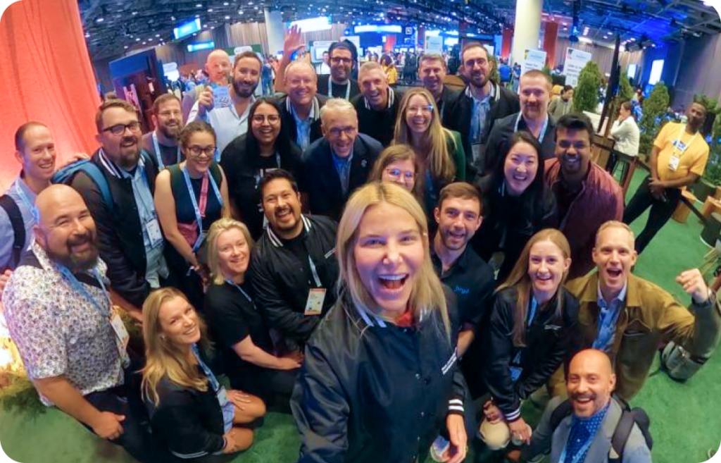 A large group of diverse individuals smiling and posing for a group selfie at an event. The background features a spacious conference venue with booths, banners, and people networking. The group appears happy and engaged, with event badges and lanyards visible on many participants.