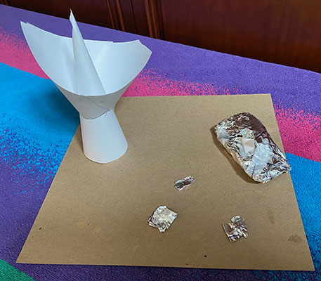 A paper model of a DSN antenna and several wrapped up foil objects on the ground next to it.