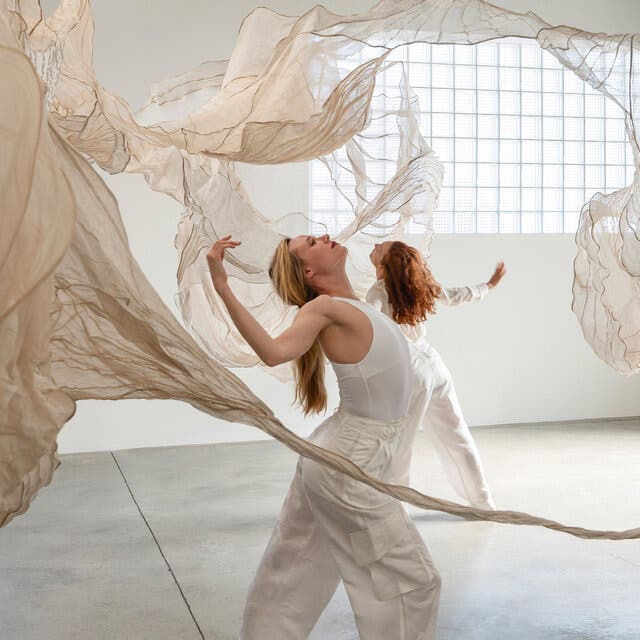 Two performers, wearing all white, dance in a gallery space surrounded by gauzy cream-colored fabrics that are seemingly suspended in mid-air.