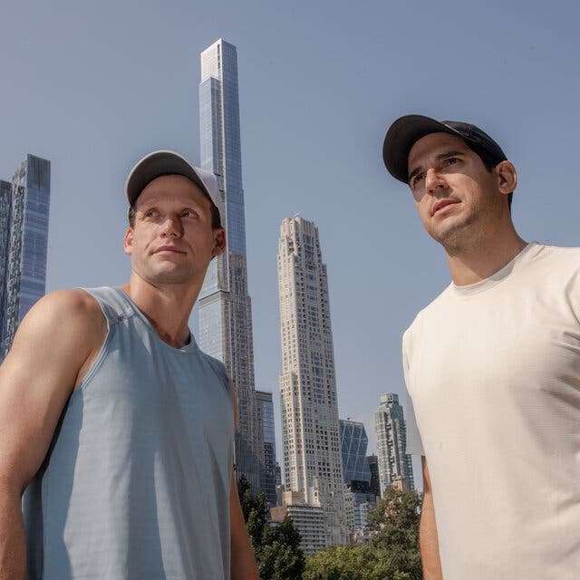 Two young men in casual shirts and baseball caps are framed by New York City skyscrapers in front of a blue sky.