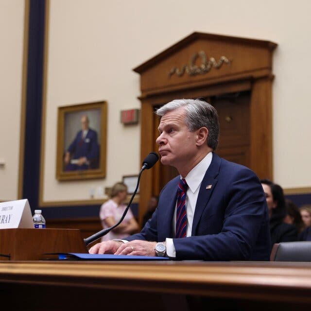 Christopher Wray, in a dark suit and a striped tie, sits at a witness table and speaks into a microphone during a House committee hearing. 