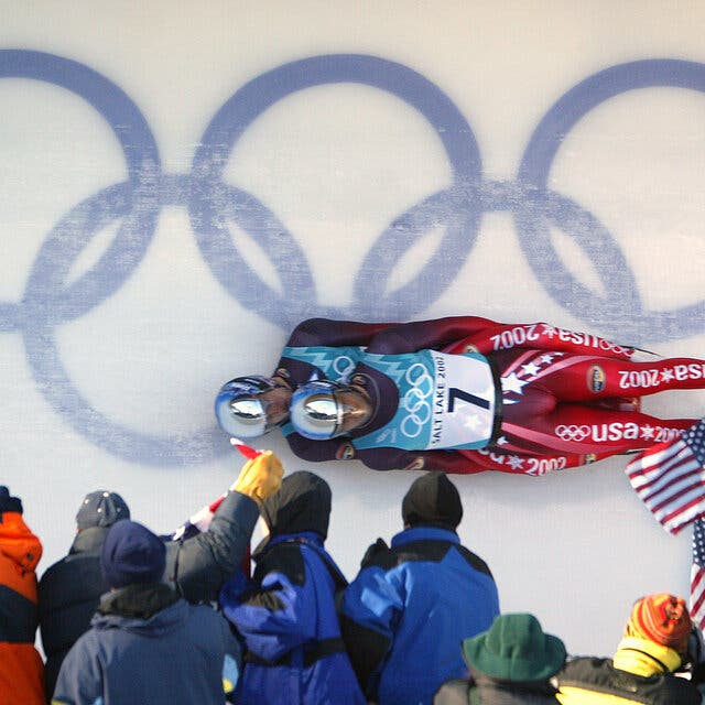A doubles luge team rounds a curve in front of fans and a blue Olympic rings logo painted under the icy track.