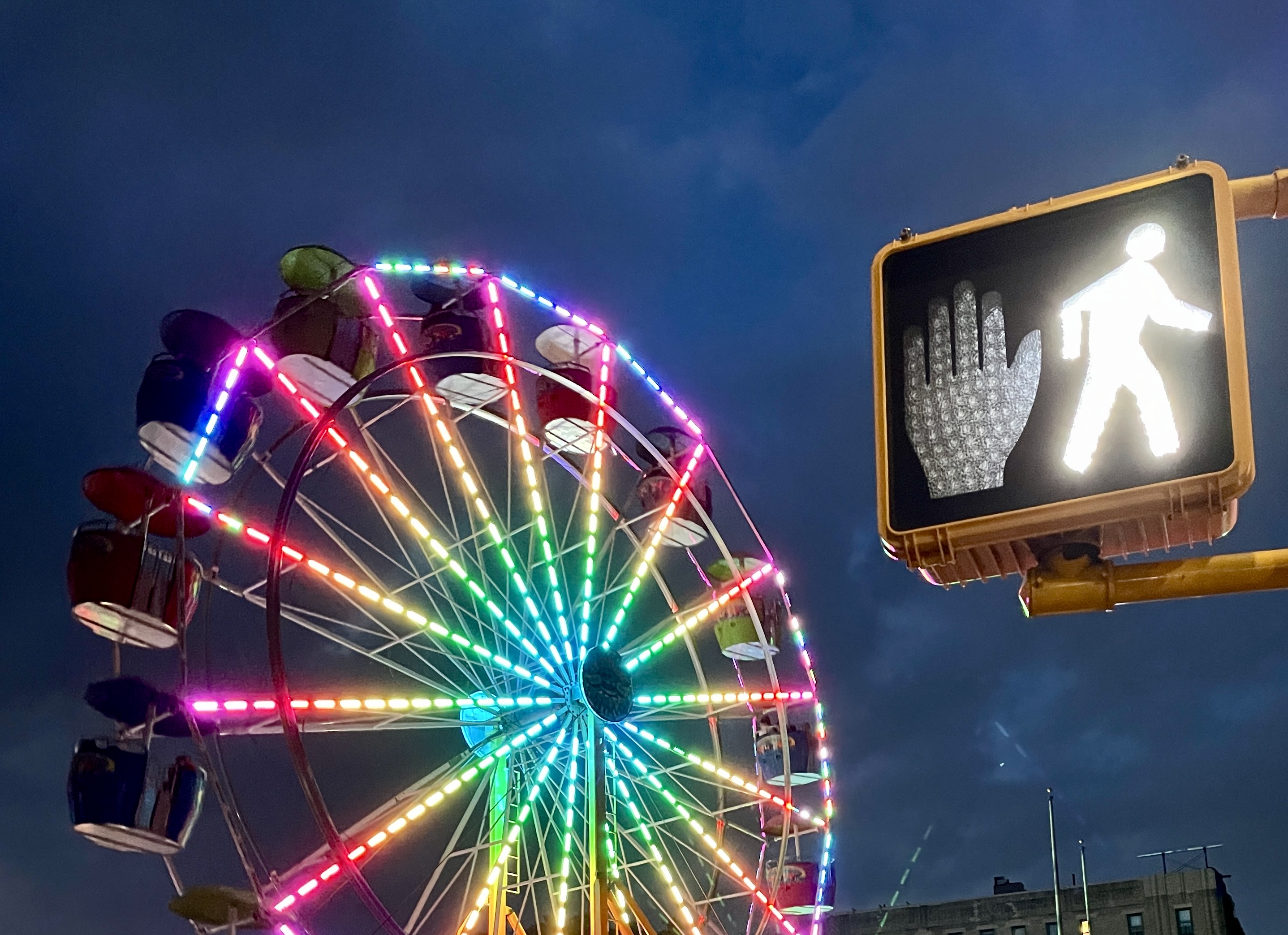 A Ferris wheel lit up in rainbow colors, with a crosswalk signal in the foreground with the walk signal illuminated.