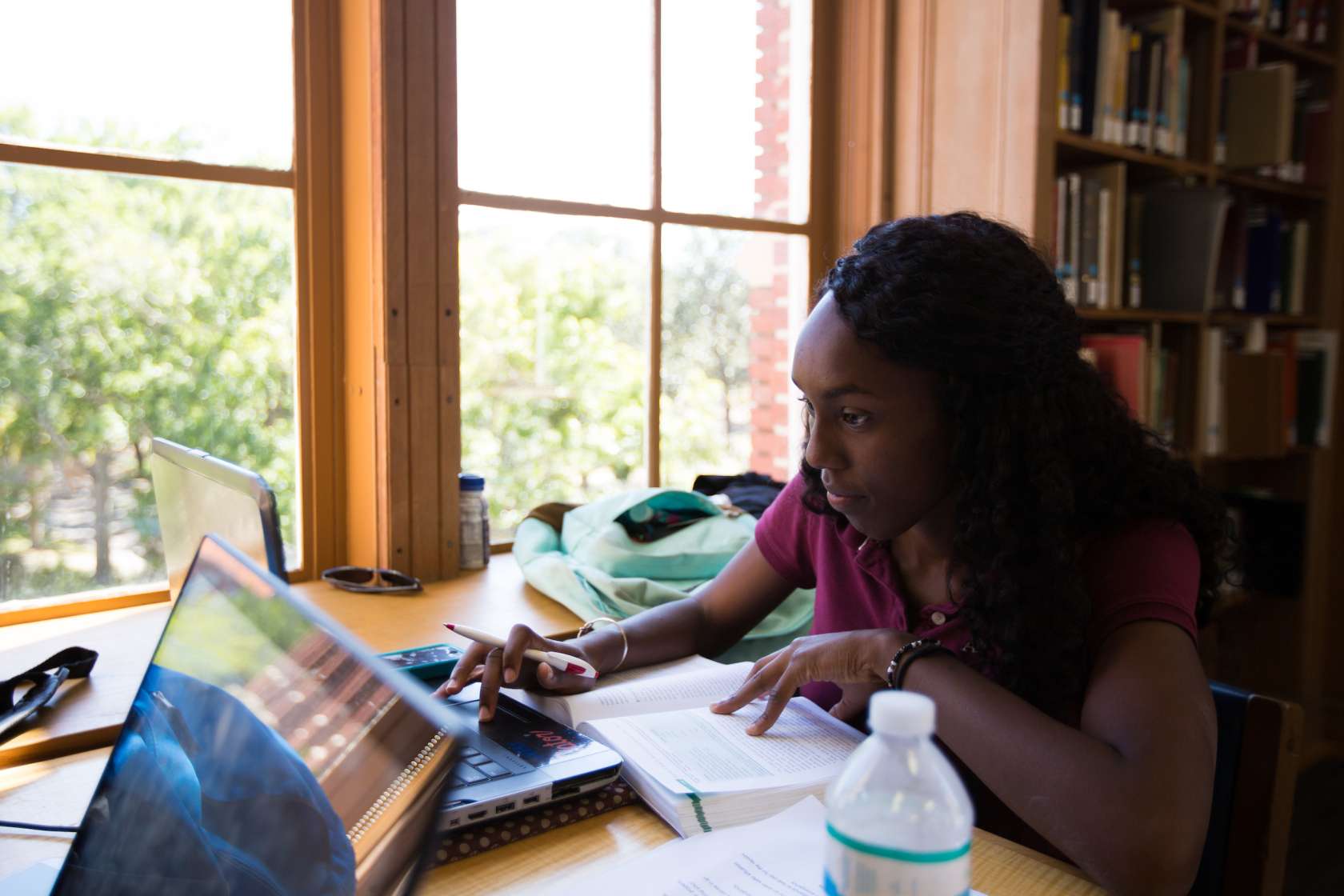 A student studies with books and laptops at a desk by a large window in the Latin American Collection.